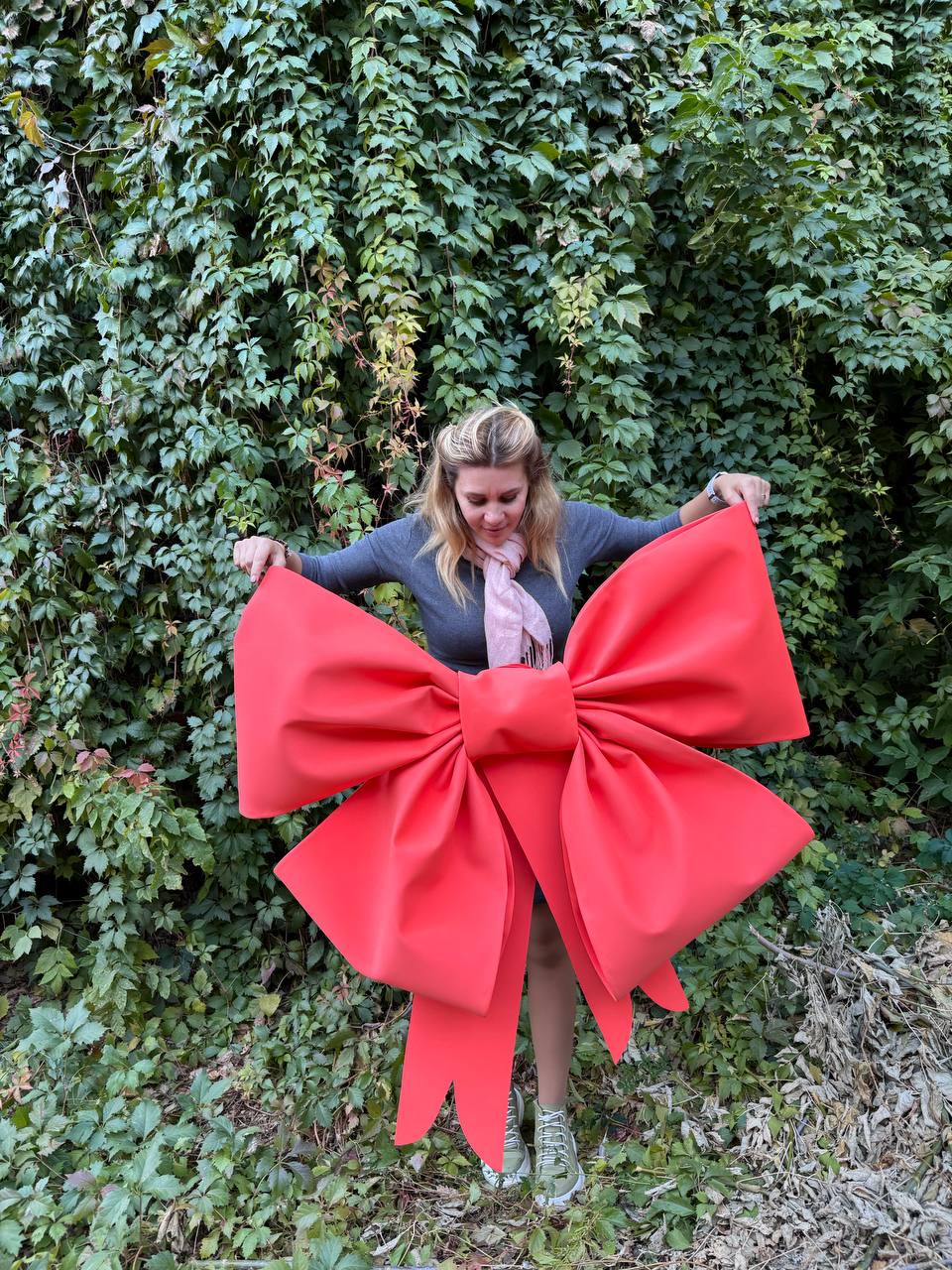 May include: A large, bright red bow with long tails. The bow is made of fabric and is being held by a person in front of a green leafy background.