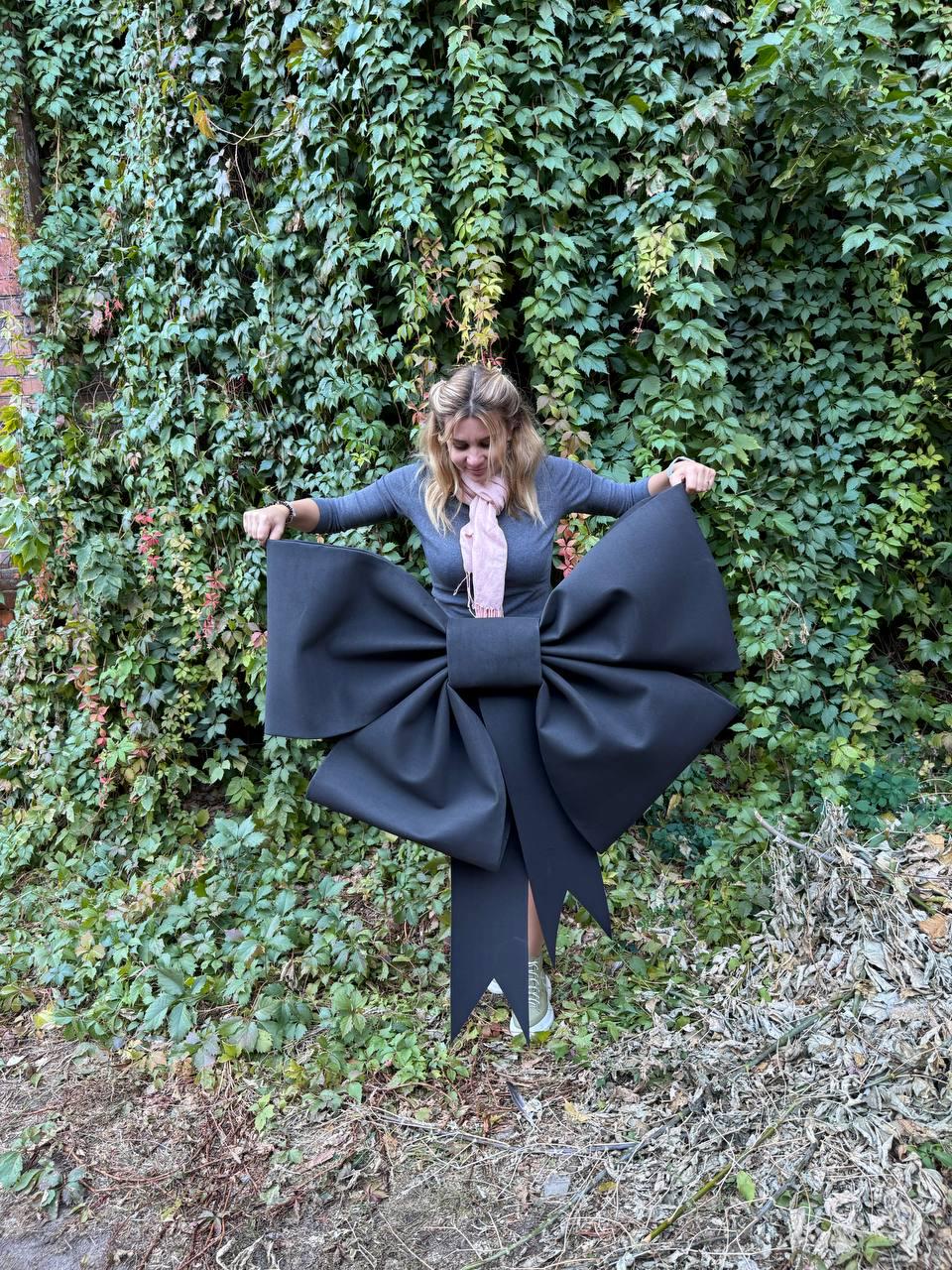 May include: A large black bow made of fabric with a long tail. The bow is being held by a person in front of a green leafy wall.