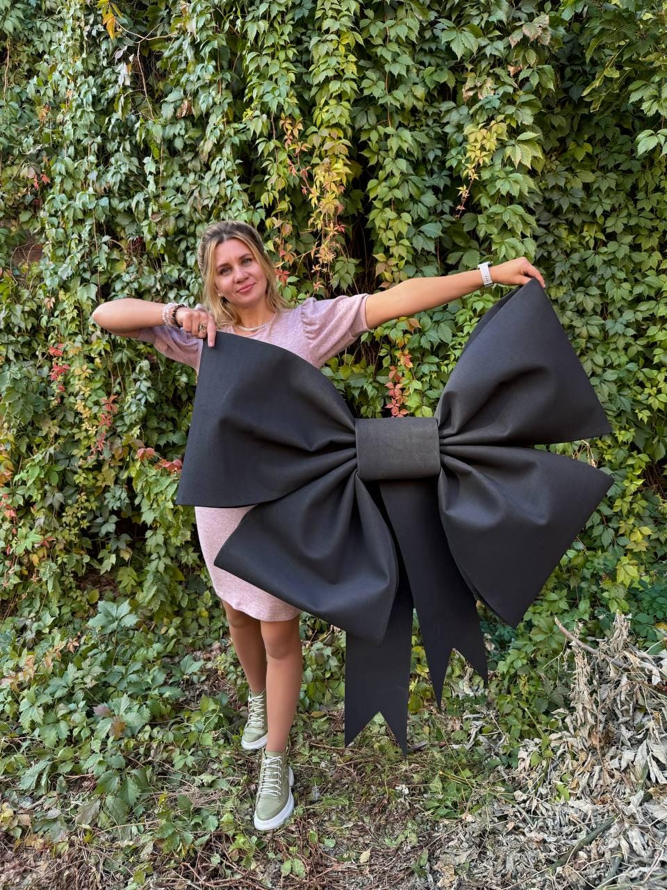 May include: A large black bow made of fabric, held by a person in front of a green leafy wall.