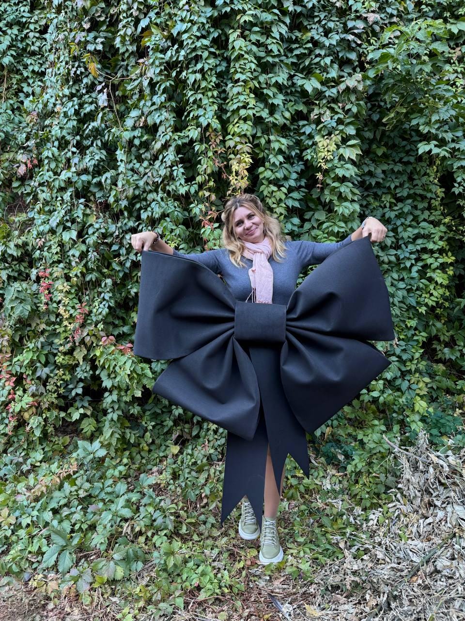 May include: A large black bow made of fabric, held by a person in front of a green ivy wall.