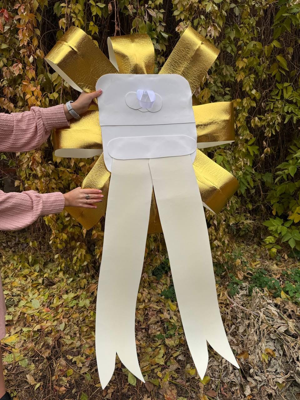 May include: A large gold bow with white satin tails. The bow is made of shiny gold material and has a white center with a small white bow.