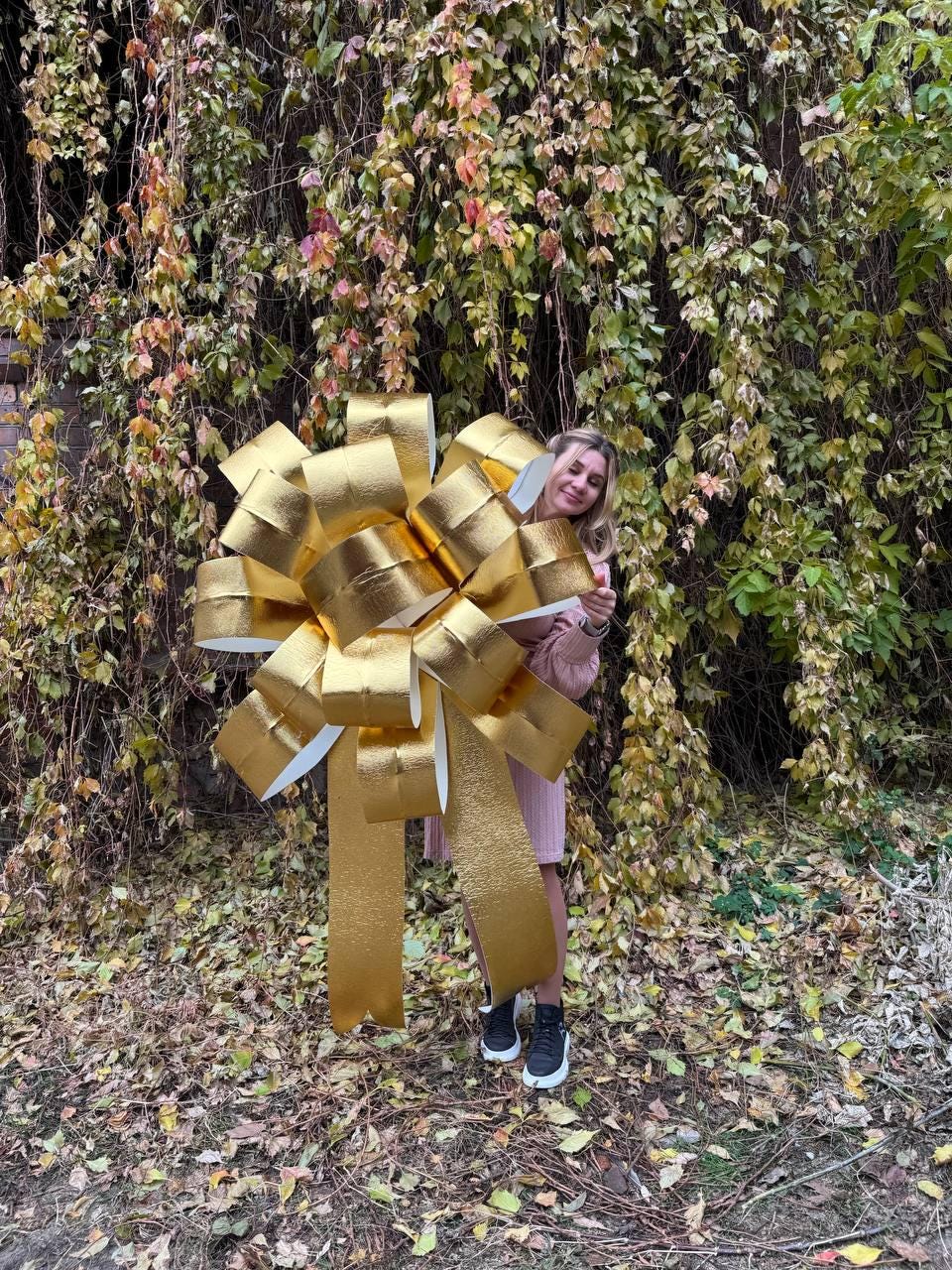 May include: A large gold bow with a white center, held by a person in front of a wall of green vines.