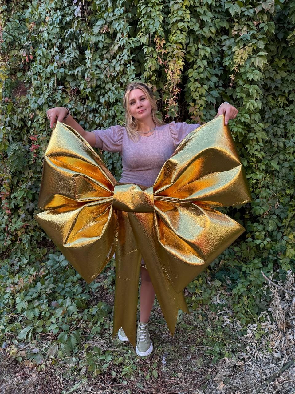 May include: A large gold bow with a person standing in front of it. The bow is made of shiny fabric and has a large, wide loop.