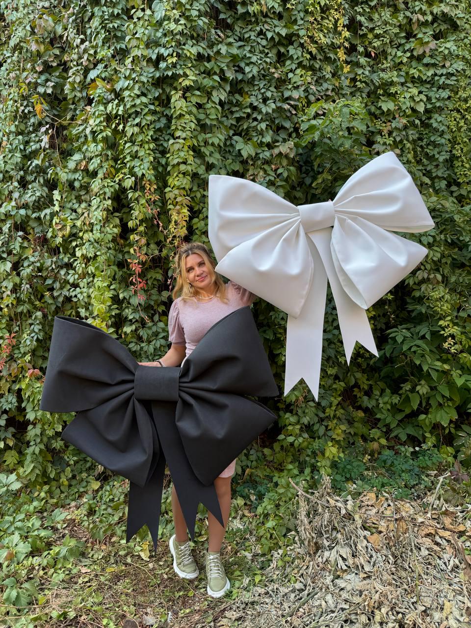 May include: Two large oversized bows, one black and one white, are displayed against a backdrop of green ivy. A person is standing between the bows.