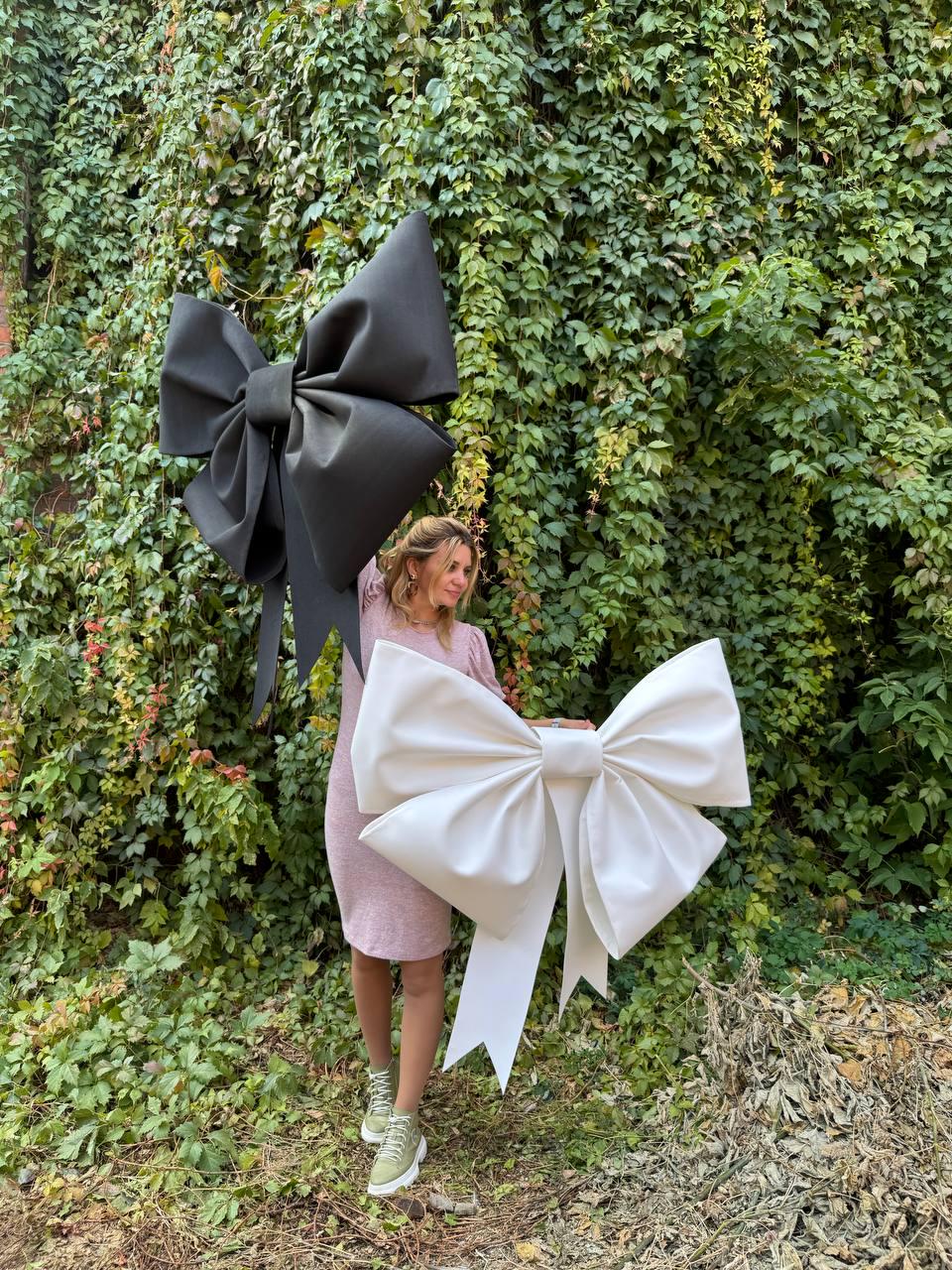 May include: A woman is holding a large white bow in front of a green leafy wall. A large black bow is behind her.