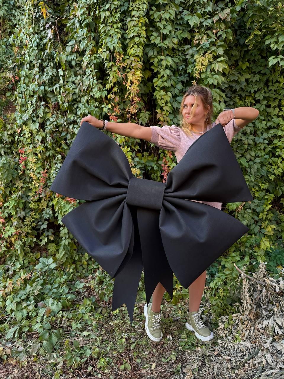 May include: A large black fabric bow with a person standing behind it. The bow is tied in a knot and has long tails. The person is wearing a pink shirt and green shoes.