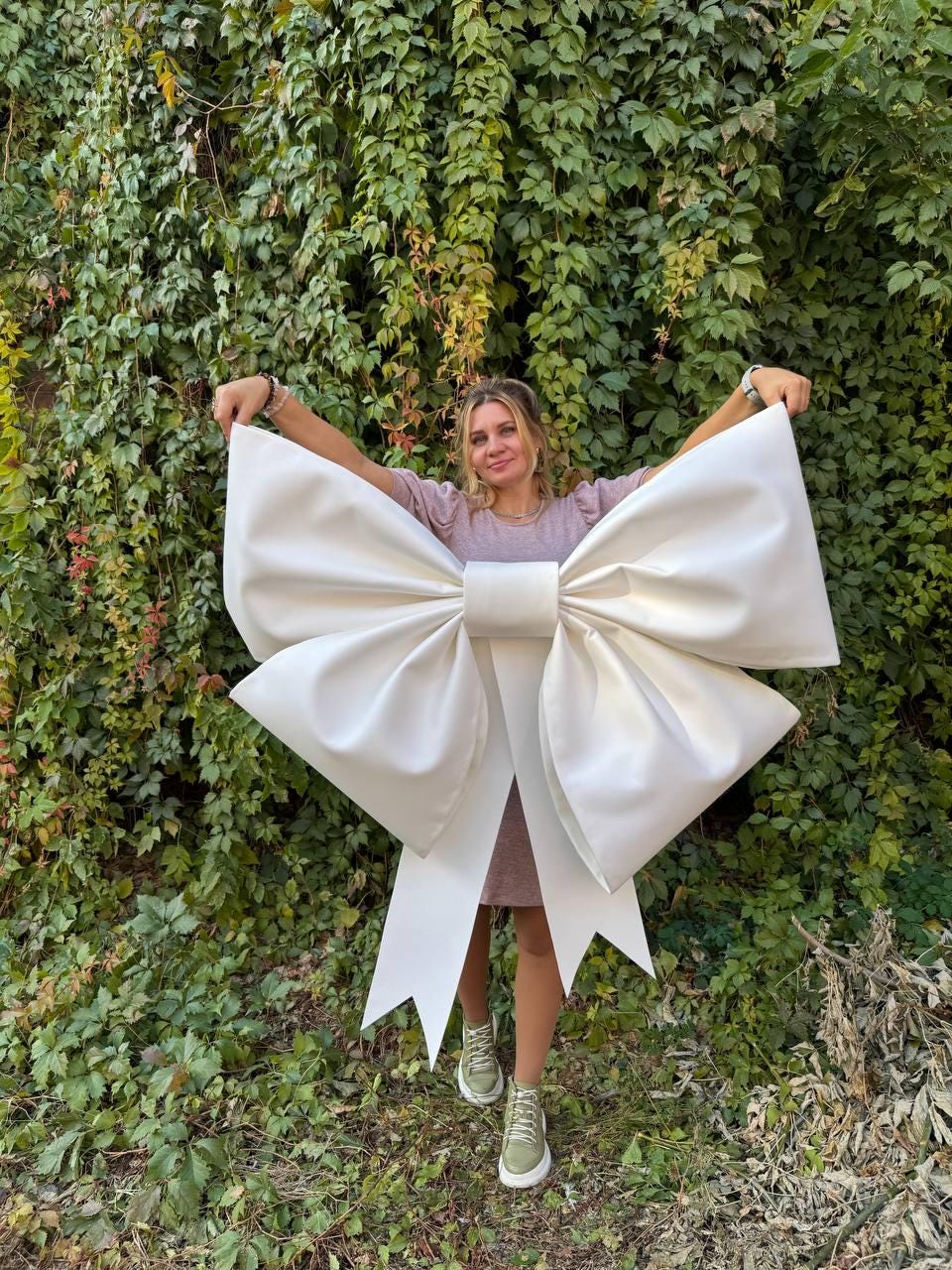 May include: A large white bow made of fabric, held by a person in front of a green leafy wall.