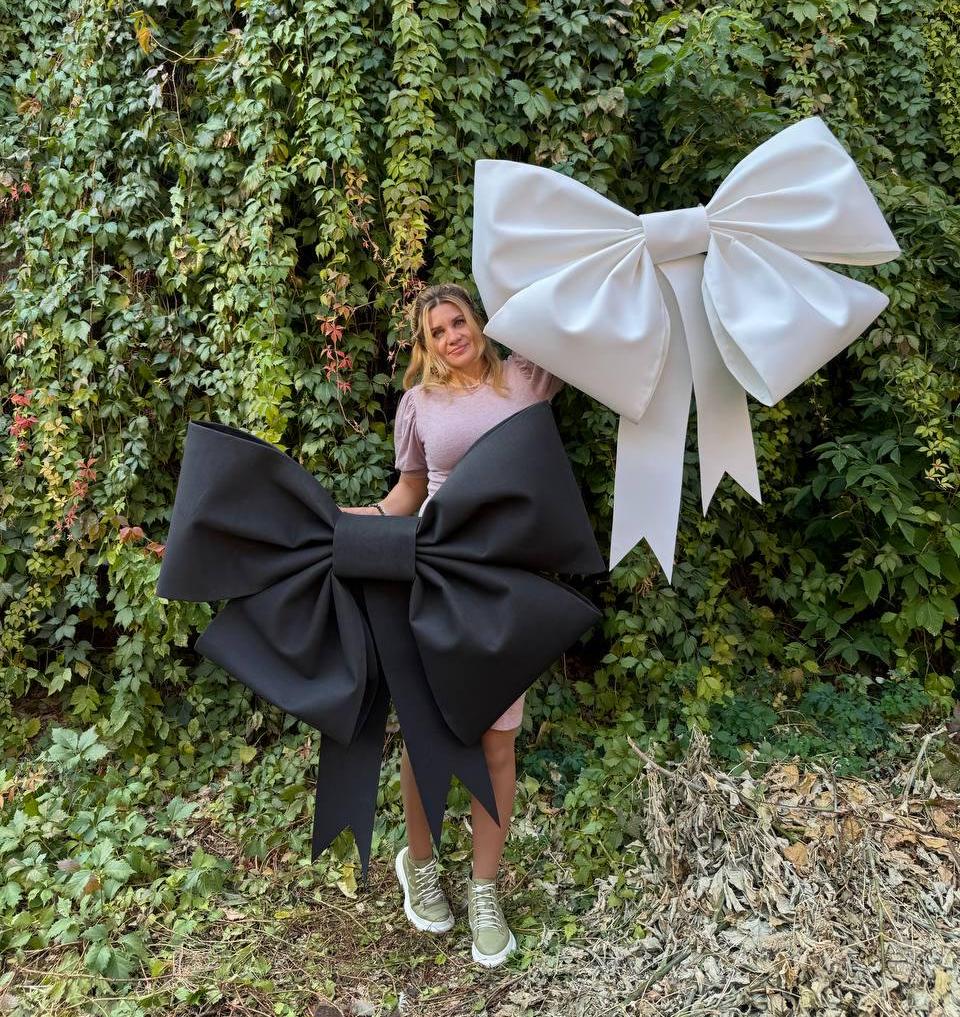 May include: A woman stands in front of a green leafy wall holding two large oversized bows, one black and one white.
