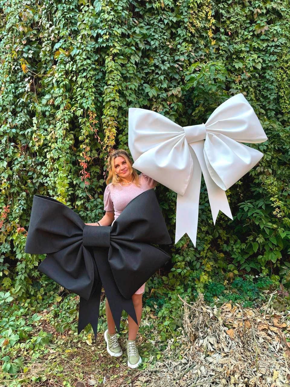 May include: Two large oversized bows, one black and one white, are displayed against a green ivy wall. A person is standing between the bows.