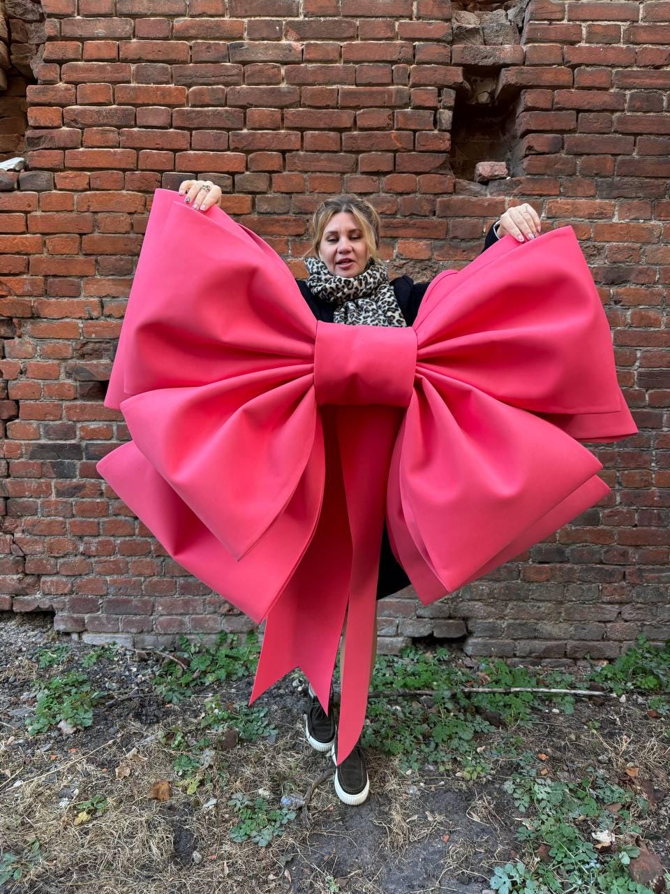 May include: A large, vibrant pink bow with long, flowing tails. The bow is held up against a weathered brick wall, showcasing its size and color. The bow's design features multiple layers and a prominent center knot.