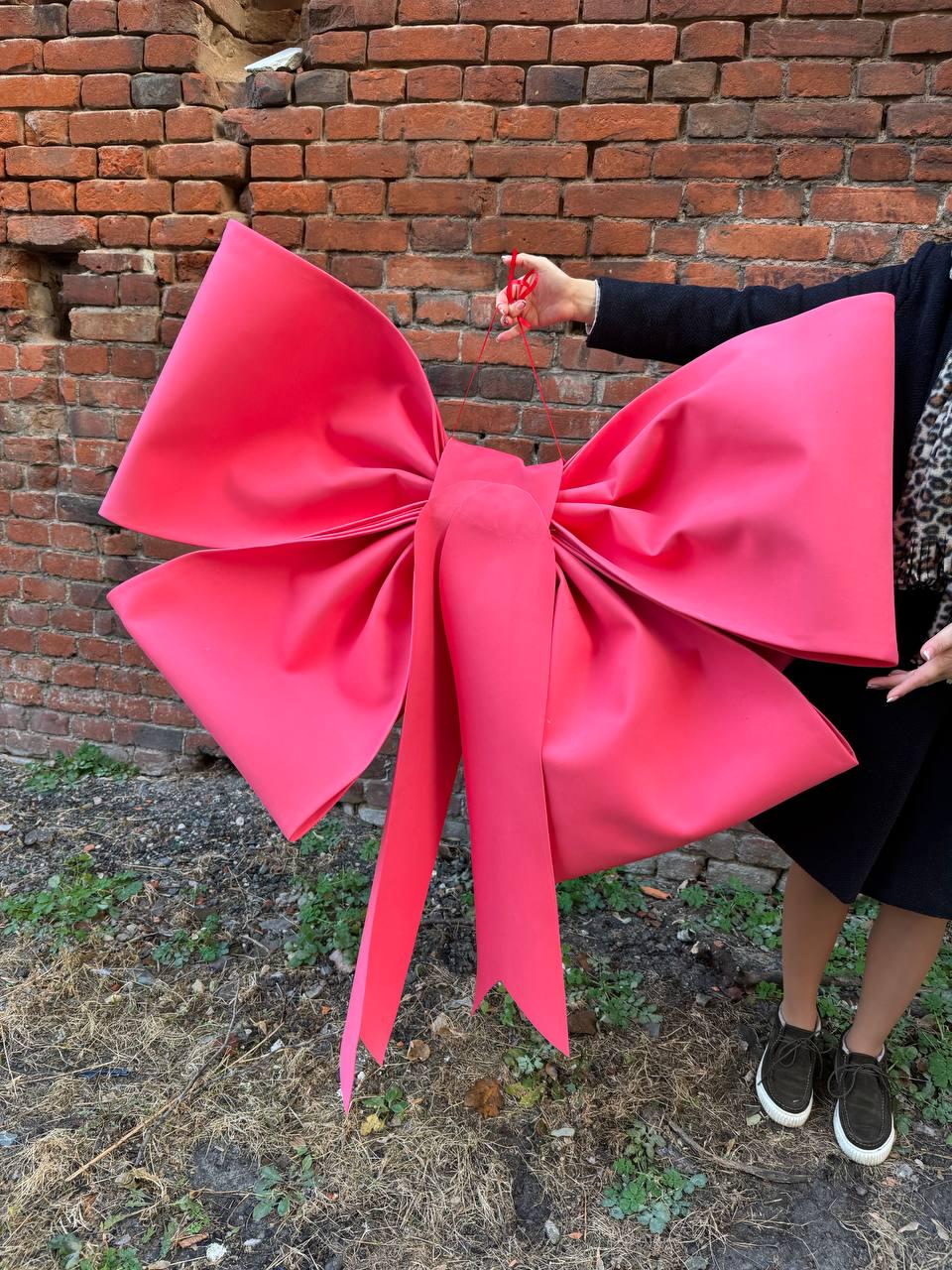 May include: A large, vibrant pink bow with long streamers. The bow is held up against a weathered brick wall. The bow's size and bright color make it a striking decorative element.