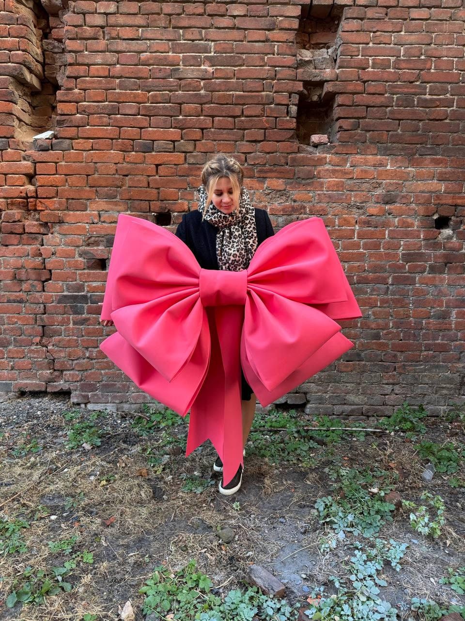 May include: A large, vibrant pink bow, possibly for decoration or a prop, dominates the image. The bow is held in front of a person, set against a weathered brick wall. The bow's size and color make it a striking visual element.