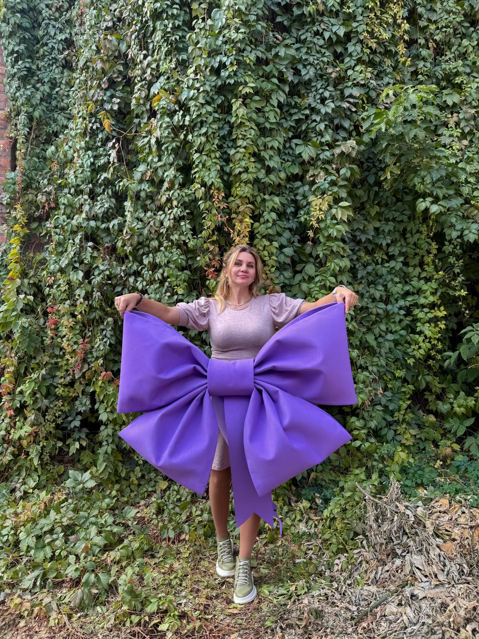 May include: A woman is standing in front of a green ivy wall holding a large purple bow. The bow is made of fabric and has a large, rounded center.
