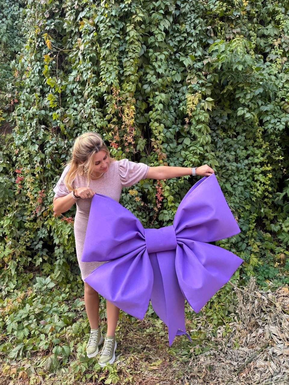 May include: A large purple bow made of fabric. The bow is being held by a person in front of a green leafy background.