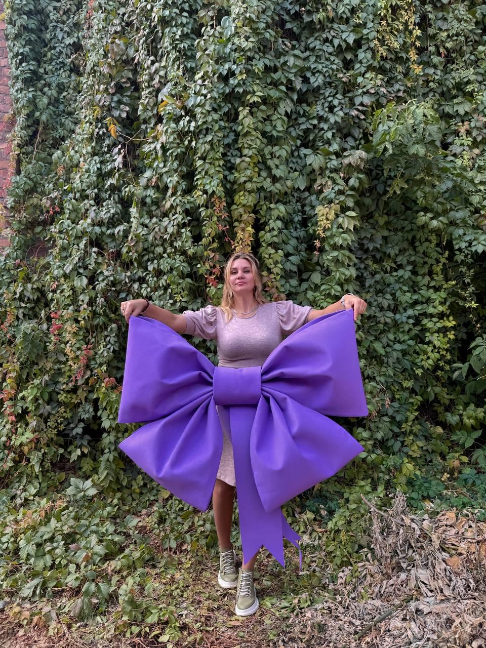 May include: A large purple bow with a long tail, held by a person in front of a green ivy wall.