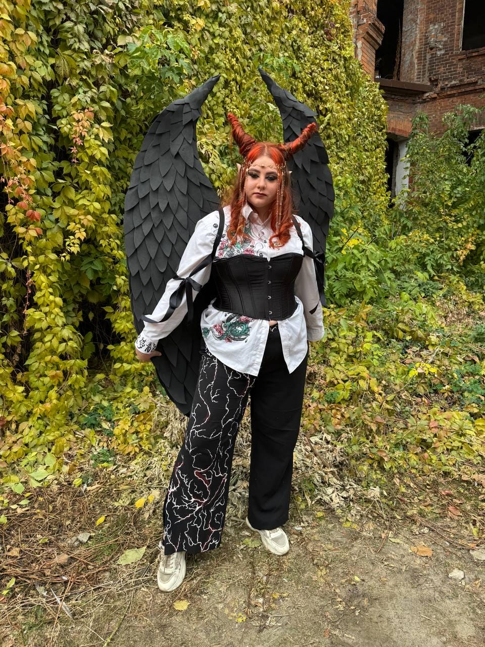 May include: A person wearing a black corset, white shirt, and black pants with a white and red design. They are wearing a pair of black devil horns and black feathered wings.