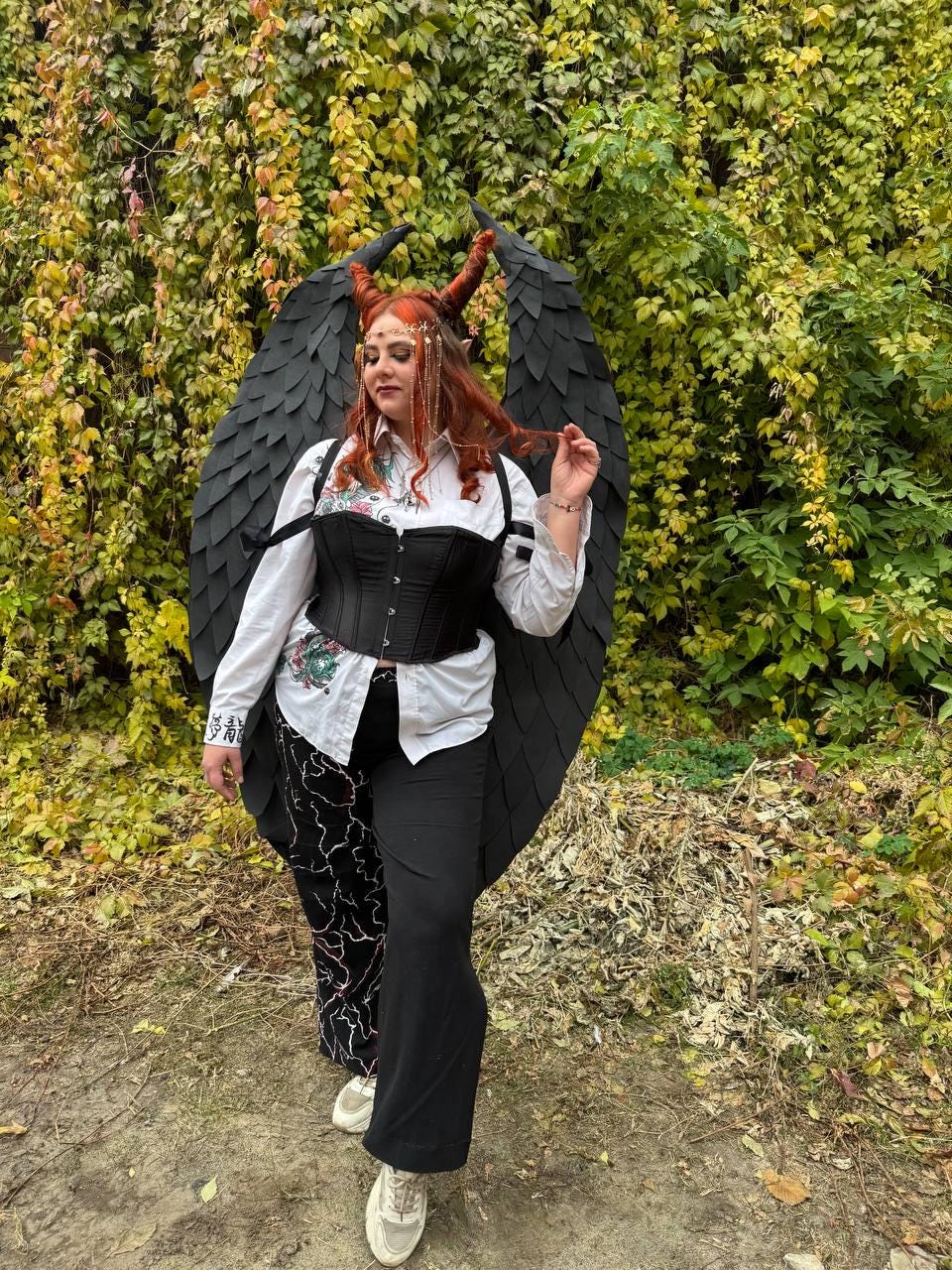 May include: A person wearing a black corset, white shirt, and black pants with a black and red lightning bolt design. They are wearing a black devil costume with large wings and red horns.