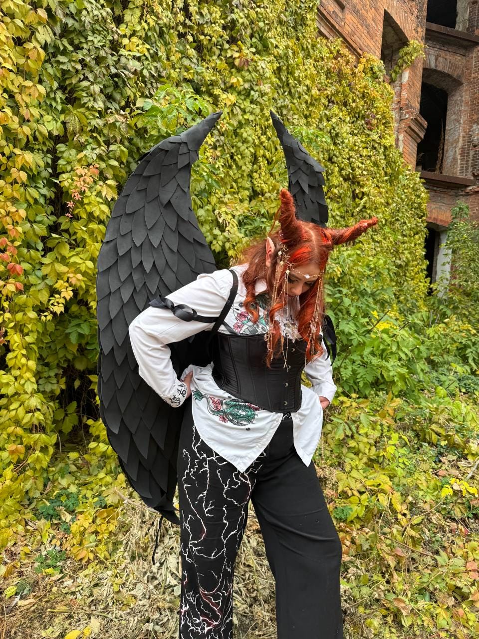 May include: A person wearing a black corset, white shirt, and black pants with a lightning bolt pattern. They are wearing a pair of black devil horns and large black wings.