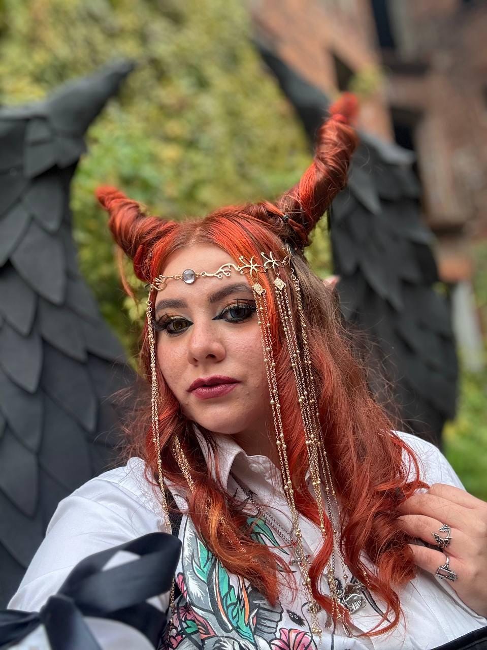 May include: A woman with red hair styled into horns wears a white shirt and a gold chain headpiece with a crystal pendant. She is wearing multiple necklaces and a ring on her finger.