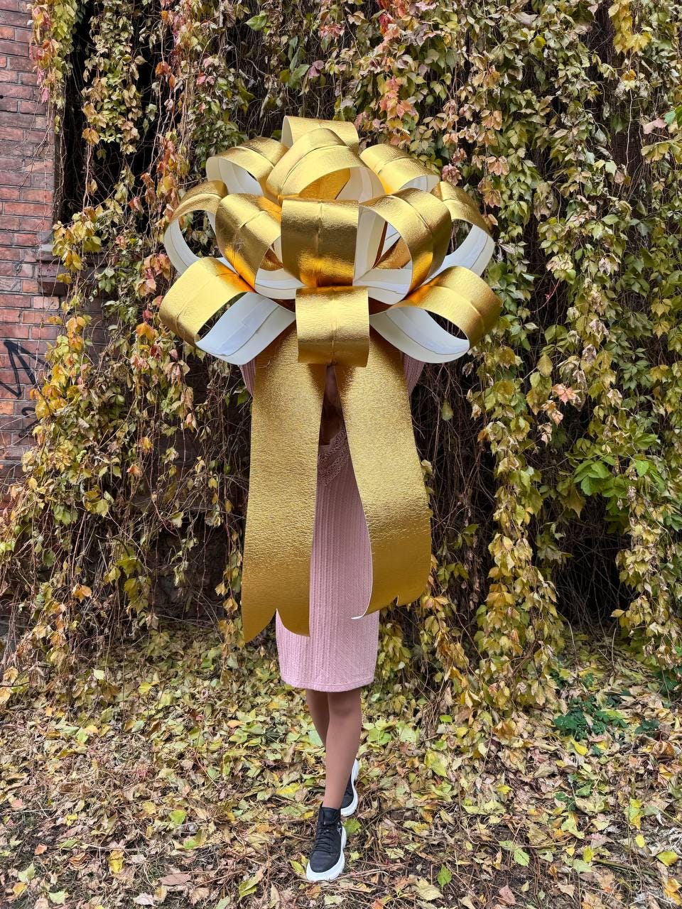 May include: A large gold bow with white accents. The bow is being held in front of a person wearing a pink sweater and black shoes.
