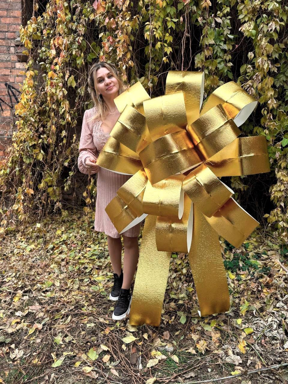 May include: A large, gold-colored bow made of shiny, crinkled paper. The bow is standing upright and is taller than the person standing next to it.