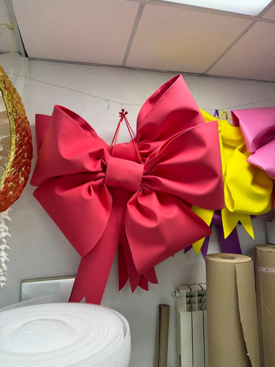 May include: A large, vibrant pink bow is the focal point, suspended against a white wall. The bow has multiple layers and a detailed design. Other colorful bows in yellow and pink are visible in the background, suggesting a craft or party supply theme.