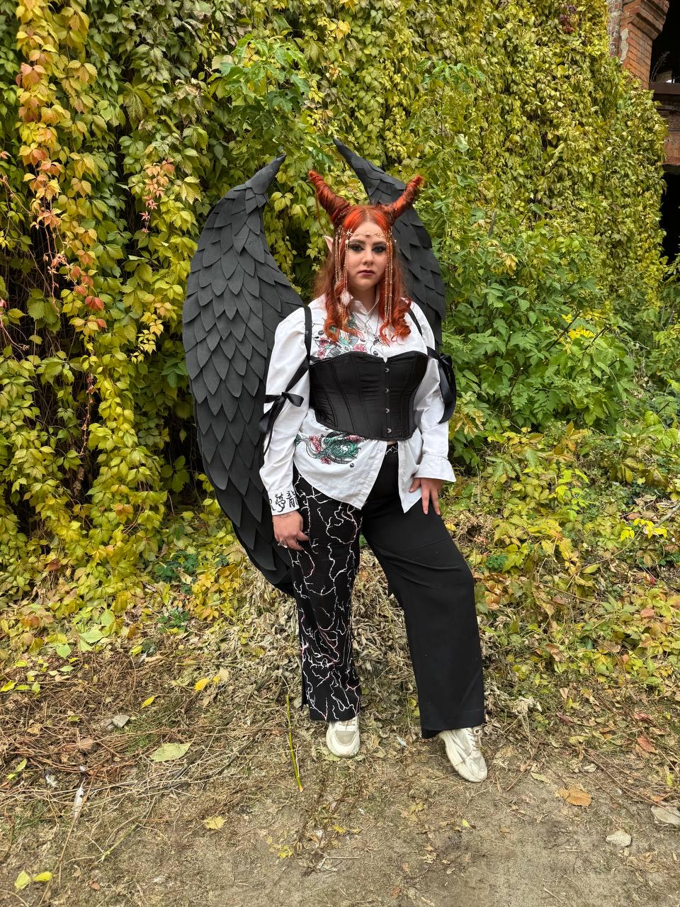 May include: A person wearing a black corset with a white shirt and black pants. They are wearing black devil horns and large black wings. The person is standing in front of a green ivy wall.