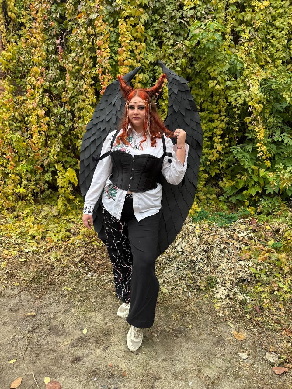 May include: A person wearing a black corset, white shirt, and black pants with a lightning bolt pattern. They are wearing a black feathered angel wing costume with red horns.