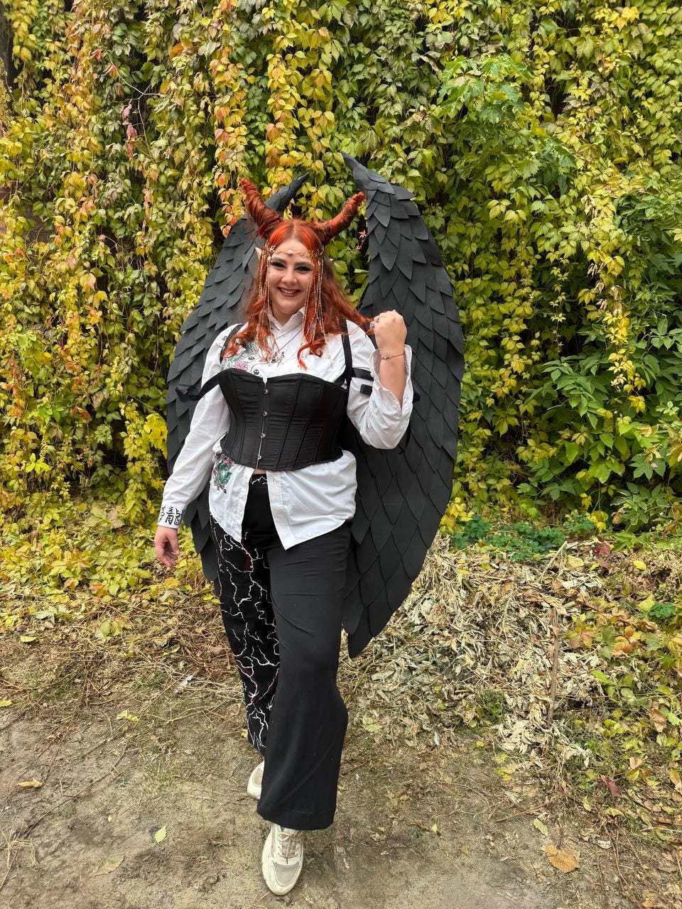 May include: A woman wearing a black corset, white shirt, and black pants with a lightning bolt pattern. She is wearing a black devil costume with horns and wings. The woman is standing in front of a green leafy background.