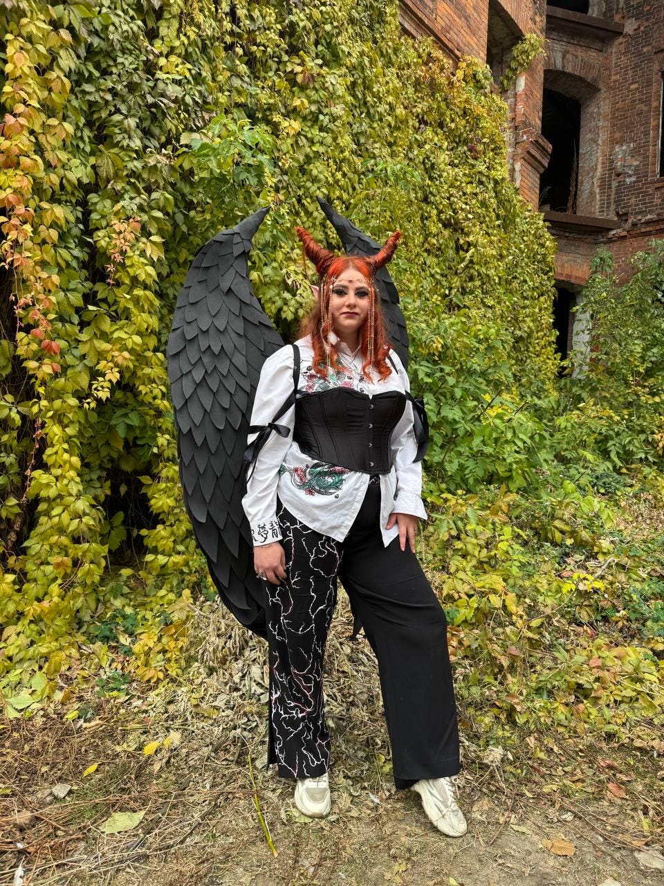 May include: A person wearing a black corset, white shirt, and black pants with a white lightning bolt pattern. They are wearing a black devil horn headband and large black wings. The person is standing in front of a brick wall covered in green ivy.