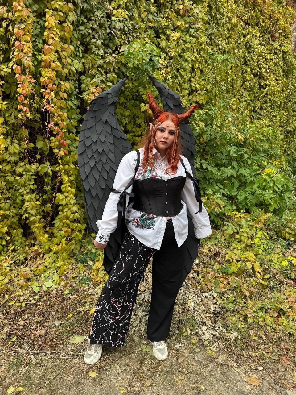 May include: A person wearing a black corset, white shirt, and black pants with a white and red design. They are wearing a black feathered wings costume and red horns. The person is standing in front of a green ivy wall.