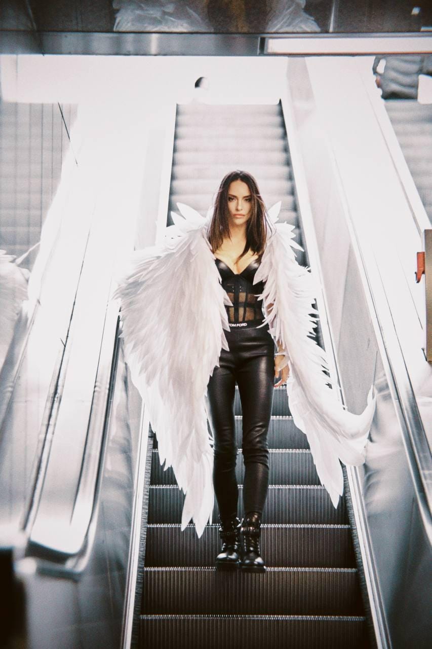 May include: A woman wearing a black leather outfit and white angel wings stands on an escalator. She is looking directly at the camera.