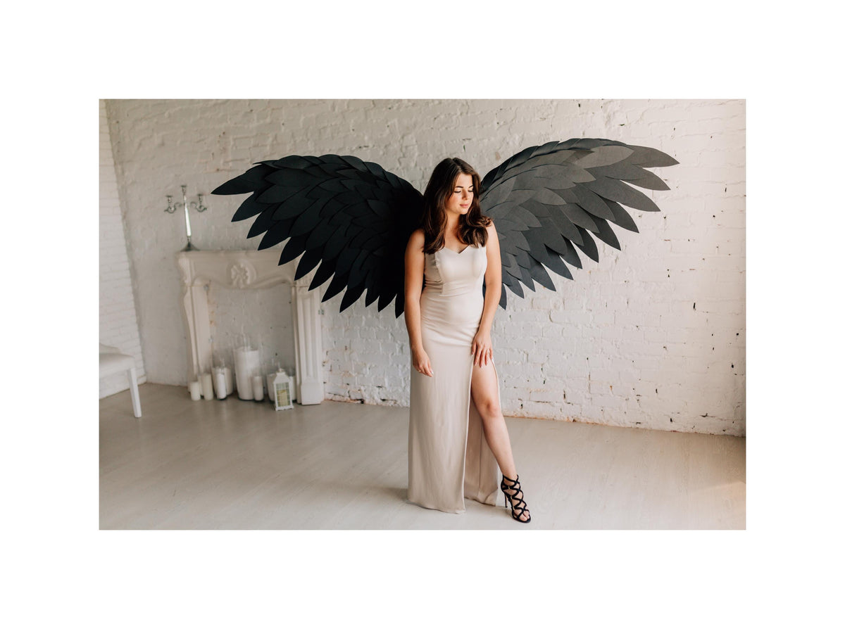 May include: A woman wearing a beige dress and black heels stands in front of a white brick wall. Large black feathered wings are mounted behind her. The room has a fireplace, candles, and a white chair.