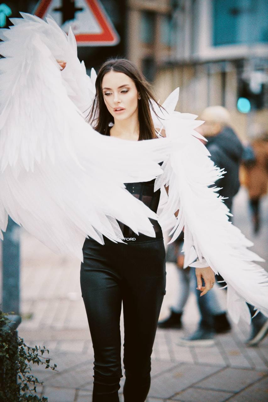 May include: A woman wearing black pants with a white waistband that says "FORD" is holding large white angel wings. The wings are feathered and have a frayed edge.