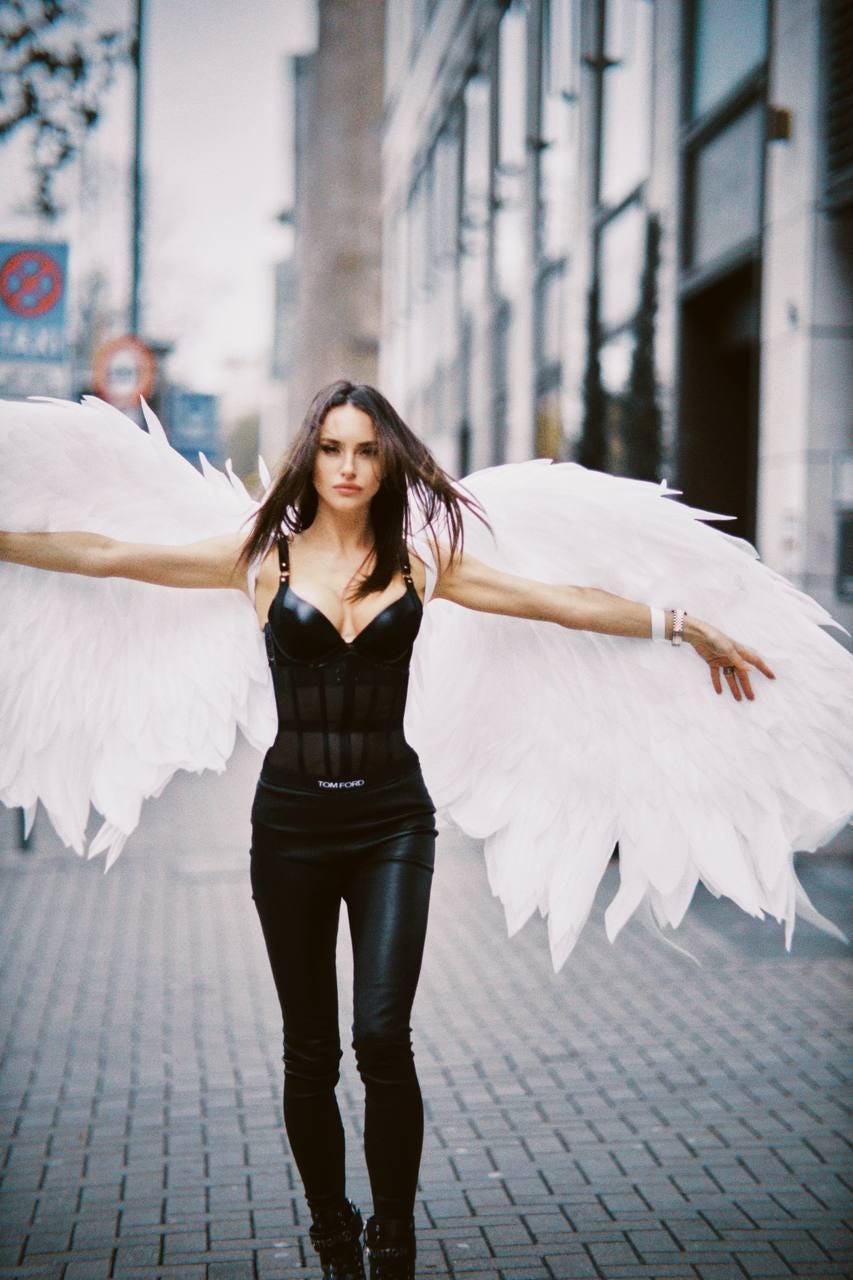 May include: A woman wearing a black leather corset with the text "TOM FORD" on the waistband, black leather pants, and white angel wings. She is standing on a brick sidewalk in front of a building.