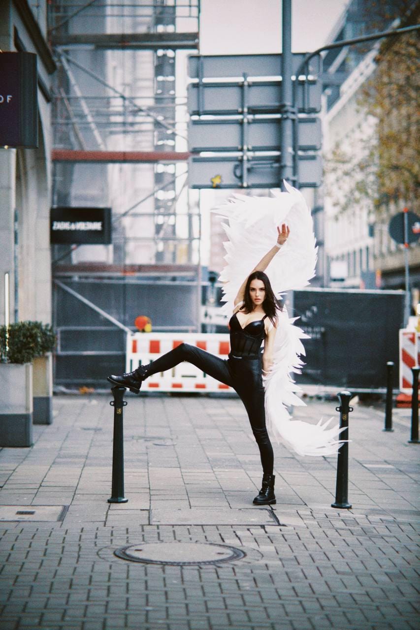 May include: A woman wearing a black corset and black pants poses with white angel wings in an urban setting. The wings are large and feathery, and the woman is looking at the camera.