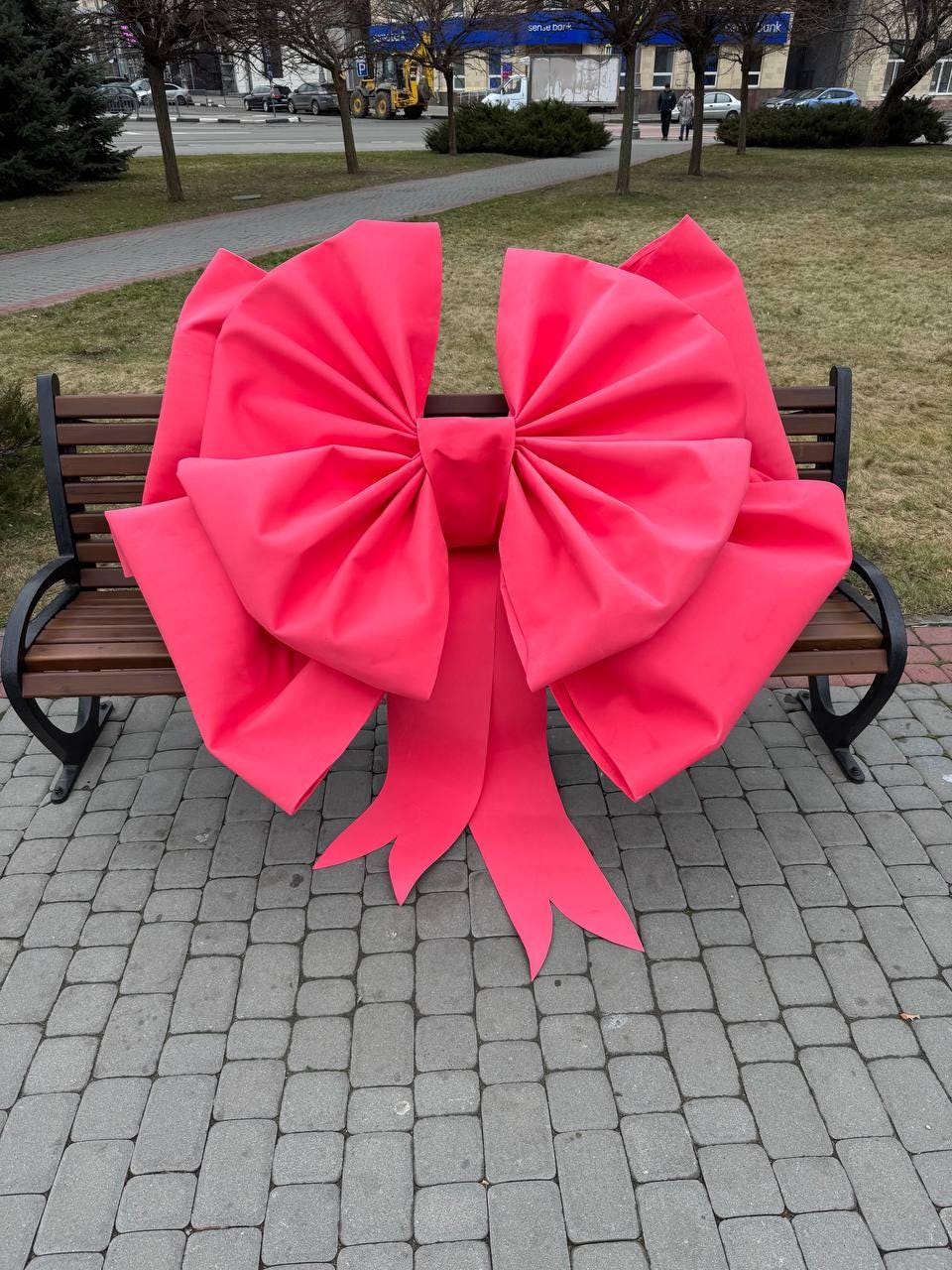 May include: A large, vibrant pink bow rests on a wooden park bench. The bow is made of a soft, pleated material, with long, flowing tails. The bow's size and color make it a striking decorative element.