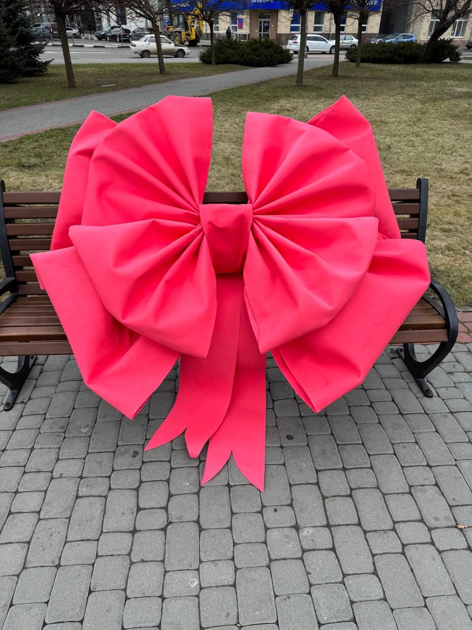 May include: A large, vibrant pink bow rests on a park bench. The bow is made of a soft, pleated fabric and features long, flowing ribbons. The bench is made of dark wood and black metal.