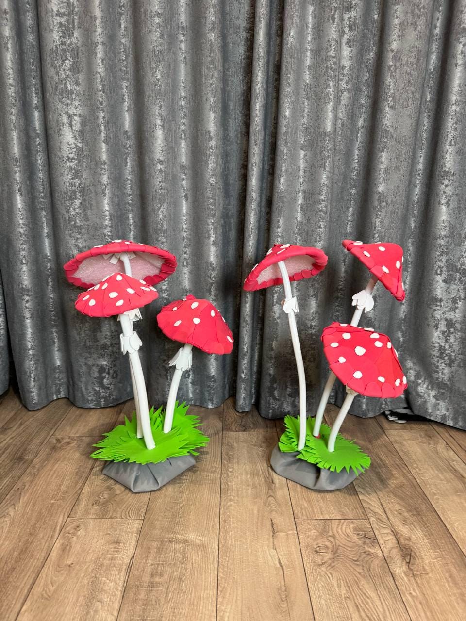 May include: A set of six red and white mushroom decorations with white stems. The mushrooms are arranged in two groups of three, with each group sitting on a green base. The mushrooms are made of paper and have a whimsical, fairy tale-like appearance.