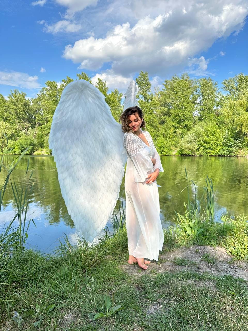 May include: A woman wearing a white dress and large white angel wings stands by a pond with green grass and reeds. The sky is blue with white clouds.