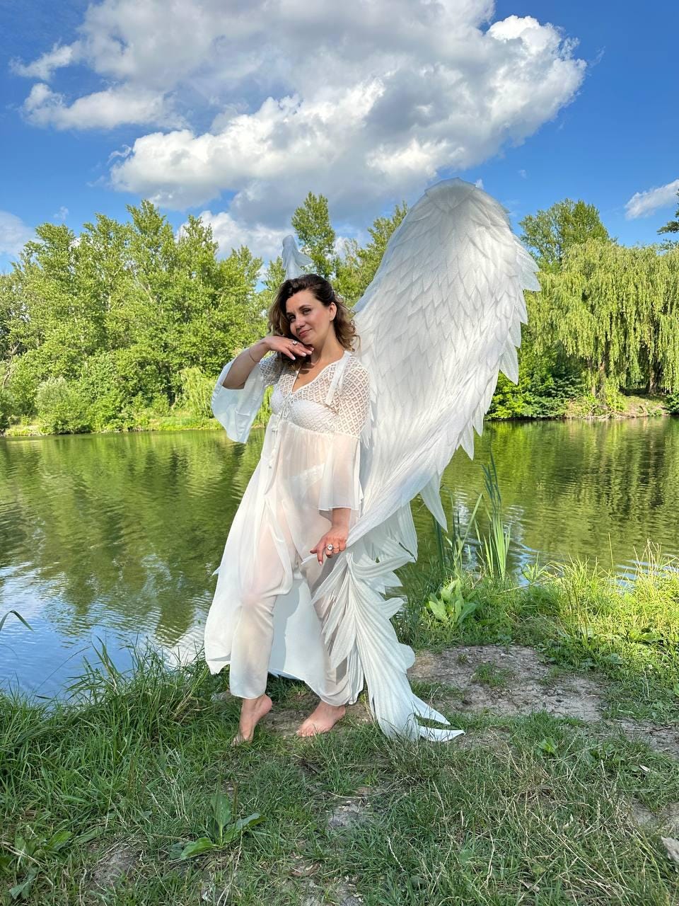 May include: A woman wearing a white dress and large white angel wings stands by a lake. The wings are made of feathers and are very detailed. The woman is smiling and looking at the camera.