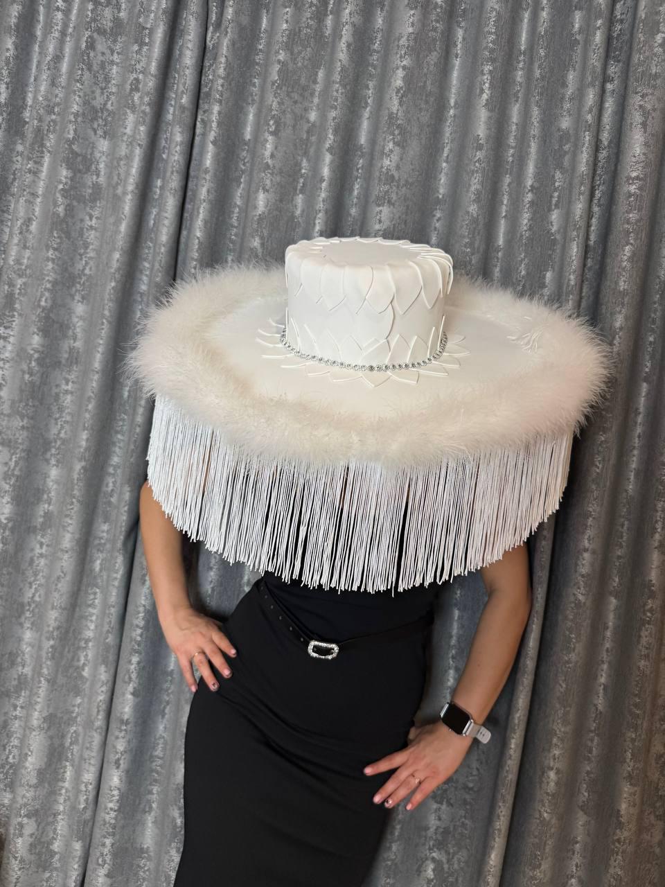 May include: A white wide-brimmed hat with a feathered trim and a white, sculpted crown. The hat is worn by a person wearing a black dress.