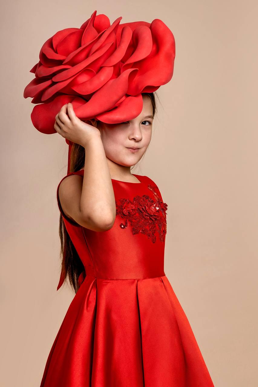 May include: A young person wearing a vibrant red dress and a large, elaborate red rose headpiece. The sleeveless dress features a fitted bodice with floral embellishments and a pleated skirt. The background is a neutral beige.