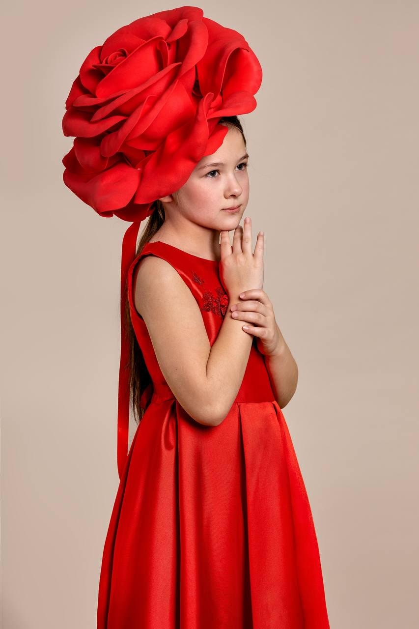 May include: A young person wearing a vibrant red dress and a large, matching rose-shaped headpiece. The sleeveless dress features a fitted bodice and a flowing skirt. The headpiece is the focal point, creating a dramatic and elegant look.