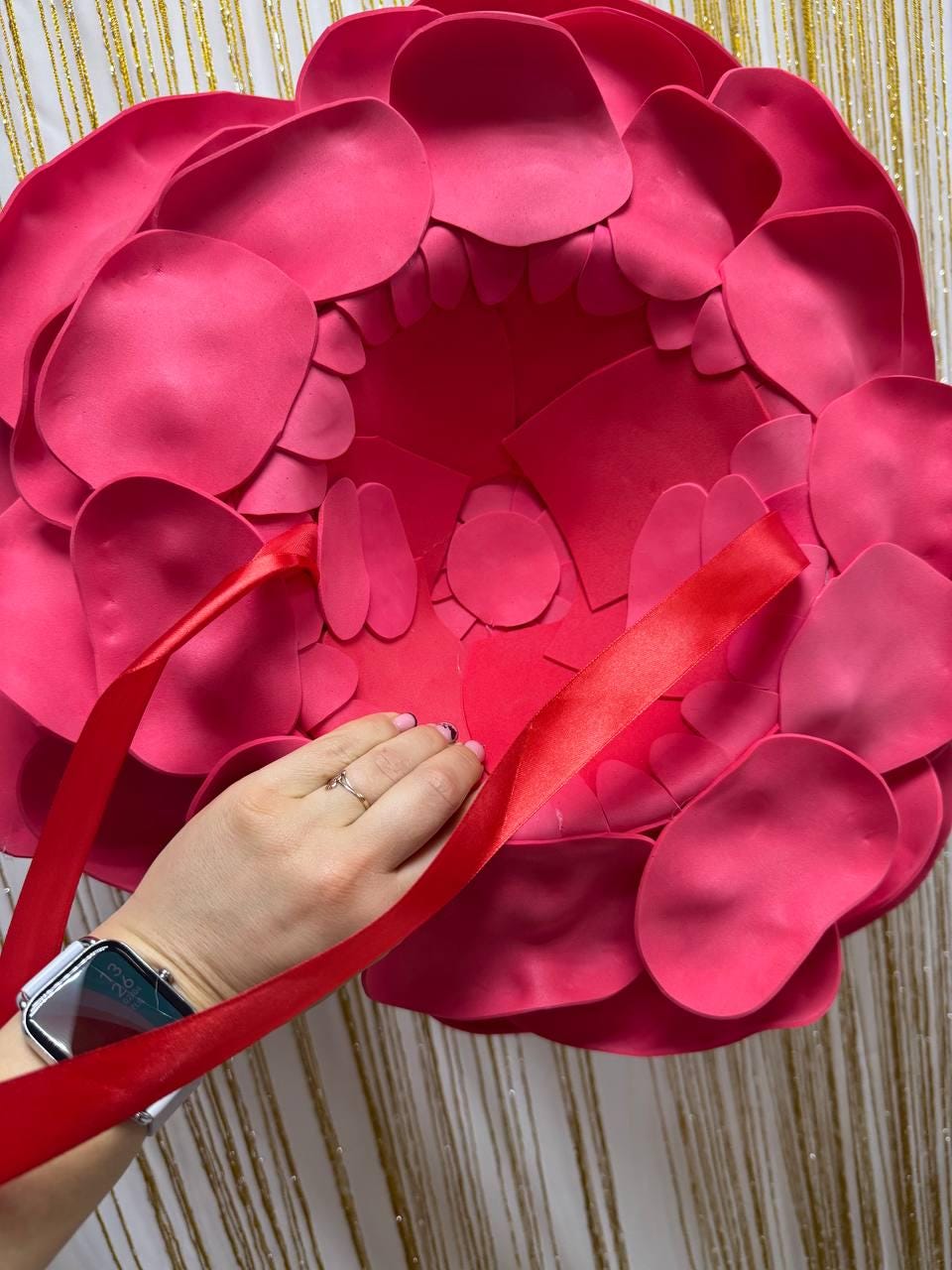 May include: A large, vibrant pink flower made of layered petals. A red ribbon is draped across the flower, and a hand with a ring is holding the ribbon. The background has a gold and white striped pattern.