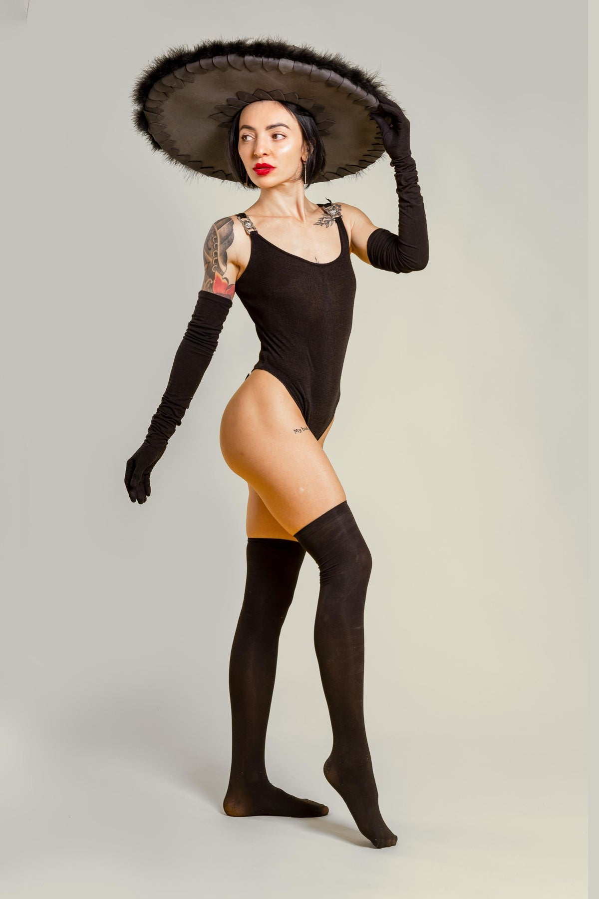 May include: A woman wearing a black bodysuit, thigh-high socks, and long gloves. She is also wearing a large, dark-colored sombrero with feather trim. The outfit is accessorized with silver shoulder straps.
