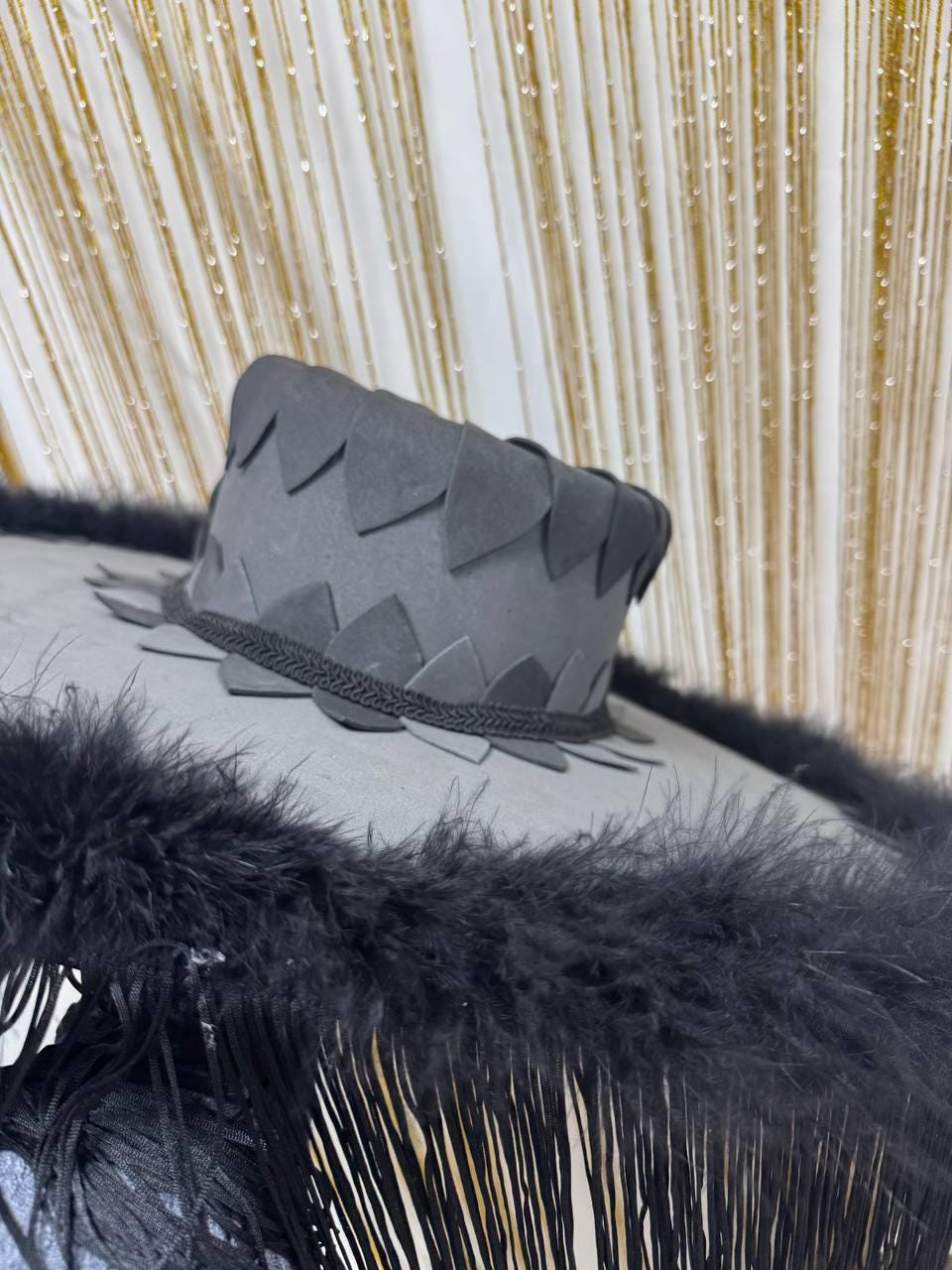 May include: A gray hat with a decorative brim and a crown with triangular cutouts. The hat is trimmed with black feathers and fringe, creating a dramatic effect. The hat is likely for a costume or special event.