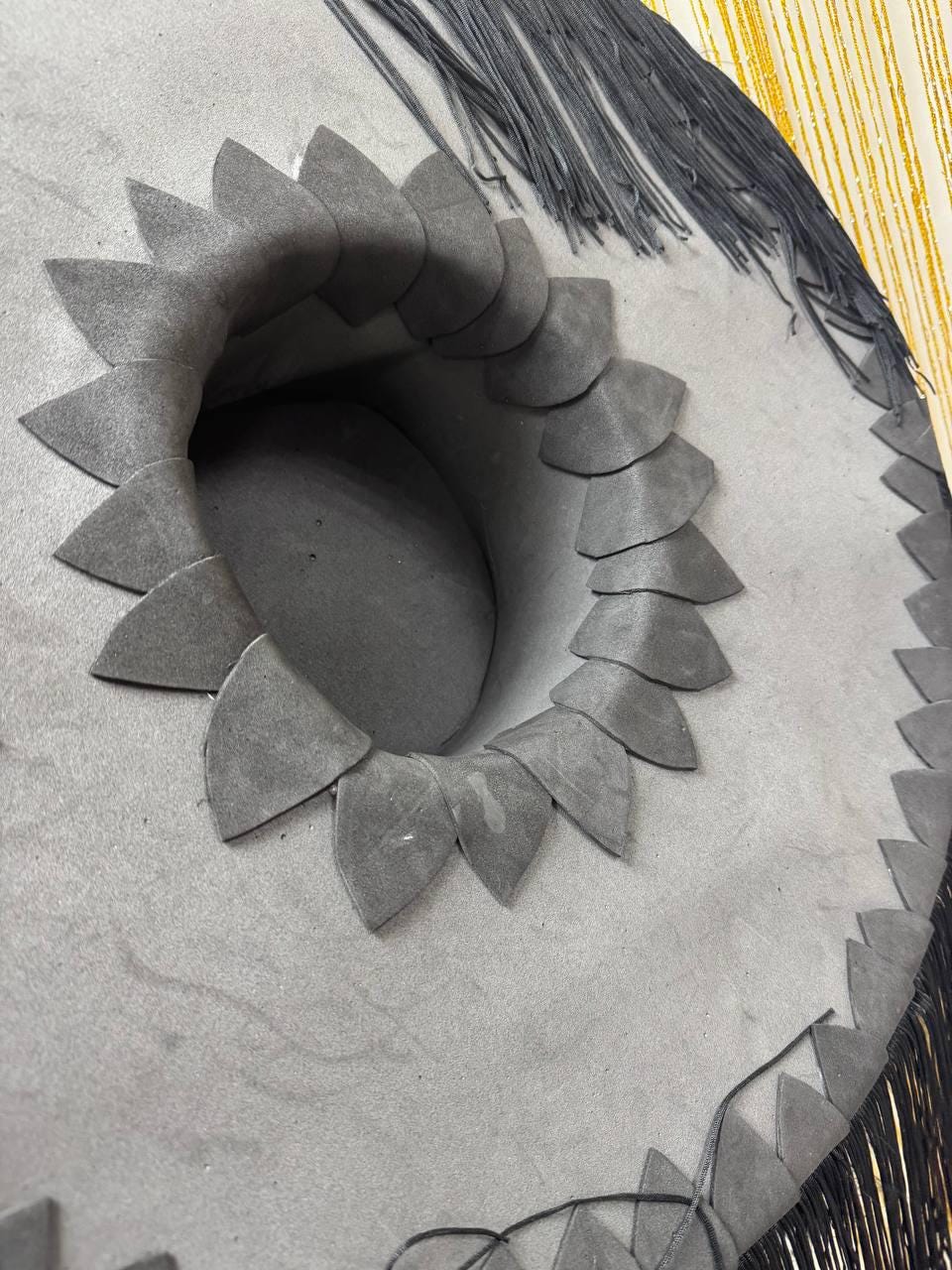 May include: A gray sombrero with a decorative brim and a central crown. The brim is adorned with a repeating pattern of petal-shaped cutouts and a fringe of dark gray tassels. The hat's design is simple yet elegant.