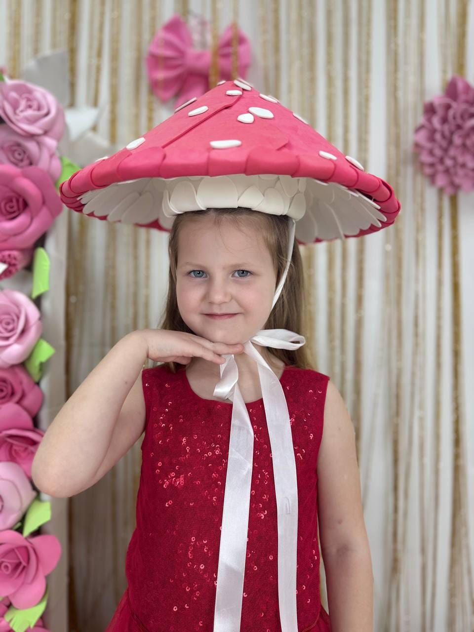 May include: A young person wearing a whimsical mushroom hat with a red cap and white spots. The hat has a white underside and is secured with a white ribbon. The person is wearing a red sequined dress. Pink paper flowers and a bow decorate the background.