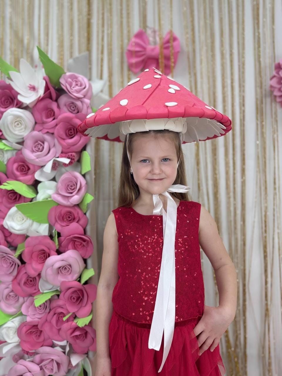 May include: A young person wearing a red sequined top and skirt, and a mushroom-shaped hat with white spots. The hat has a white trim and is secured with a headband. A large floral arrangement of pink and white paper roses is in the background.